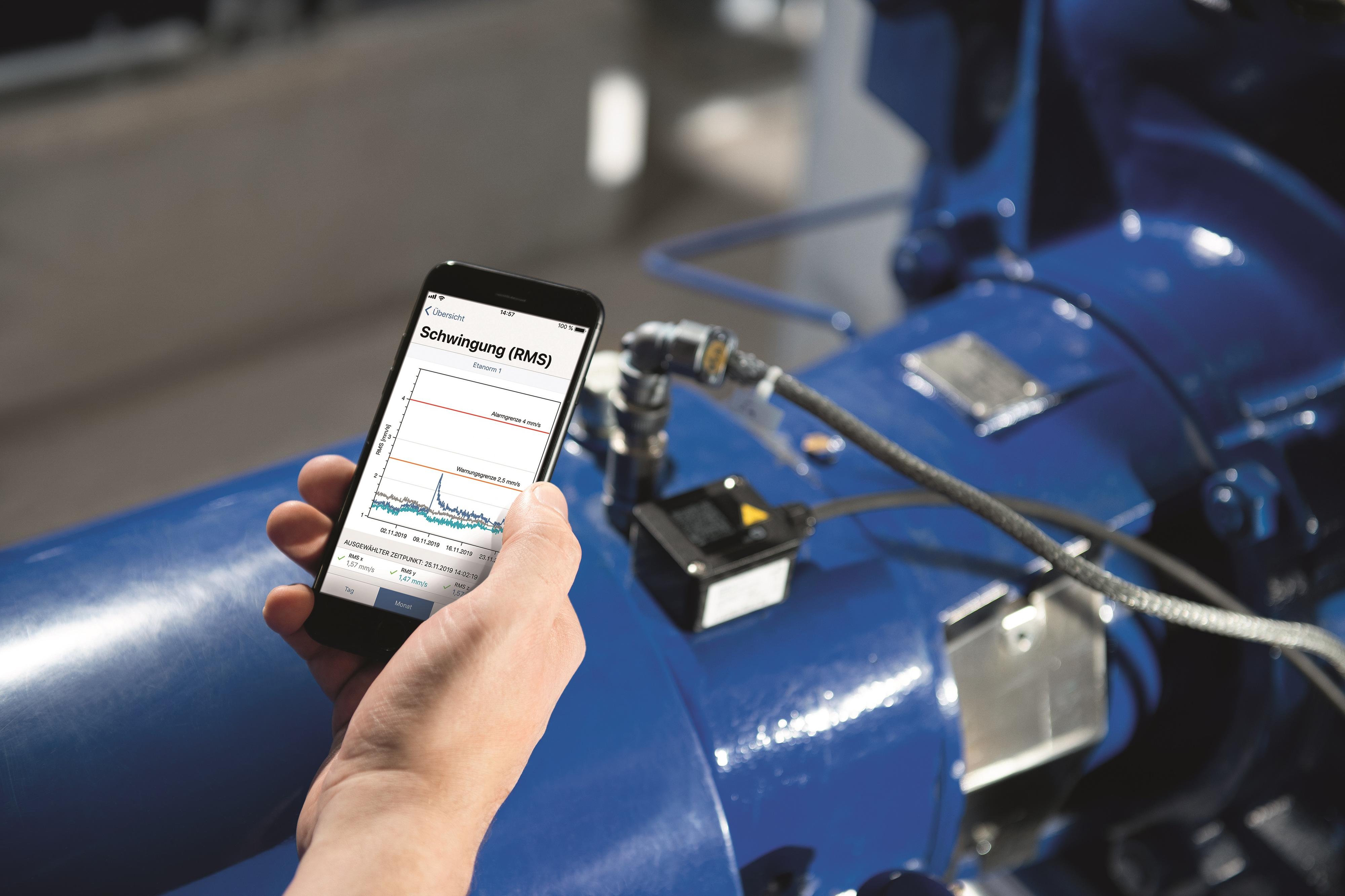 Main tenant un téléphone portable avec un graphique affiché à l'écran. En arrière-plan, une pompe KSB avec le KSB Guard installé.